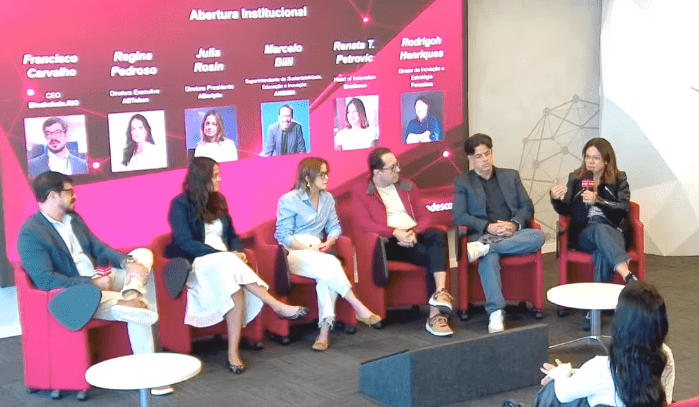 Un panel de seis expertos en finanzas y tecnología debaten sentados en sillones rojos frente a una pantalla interactiva con sus nombres. Entre ellos se encuentra Renata Petrovic, jefa de innovación de Bradesco. El evento está centrado en la apertura institucional y la tecnología blockchain en Brasil.